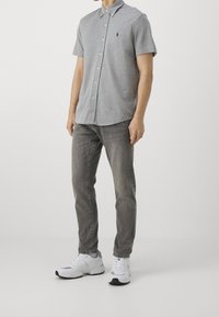 Camicia grigia a maniche corte con colletto e bottoni, abbinata a jeans grigi e sneakers bianche da ginnastica. Presenta un piccolo logo blu navy sul petto.