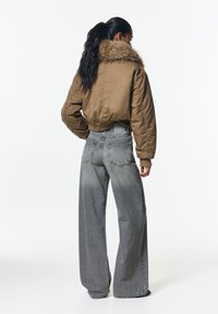 Jachetă bomber maro cu guler din blană sintetică, asortată cu blugi gri, cu picioare largi. Prezintă manșete cu model energe și buzunare la spate pe blugi.