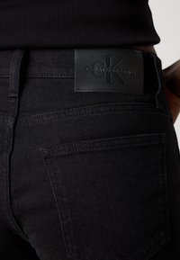 Black denim jeans with a leather Calvin Klein Jeans patch on the waist. Features include stitching details and back pockets.