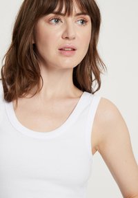 Biała, prążkowana koszulka na ramiączkach z okrągłym dekoltem i szerokimi ramiączkami, o gładkiej fakturze i dopasowanym kroju, noszona przez modelkę.