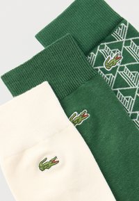 Τρία ζευγάρια κάλτσες σε λευκούς και πράσινους τόνους, το καθένα με κεντημένο πράσινο λογότυπο κροκόδειλου, ένα ζευγάρι με γεωμετρικό σχέδιο.
