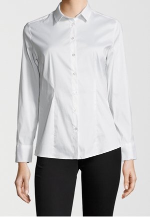 Femme portant une chemise blanche cintrée à manches longues avec boutons et un pantalon noir, sur un fond clair uni.