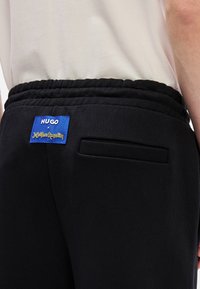 Μαύρο παντελόνι τζόκινγκ με υφή, ελαστική μέση με πλεχτό σχέδιο και μπλε ετικέτα μάρκας με κίτρινο κείμενο στο πίσω μέρος.