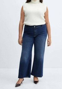 Vrouw draagt een mouwloze witte coltrui, hoogtaillotte wijde blauwe jeans en zwarte pumps met puntige neus, tegen een effen achtergrond.