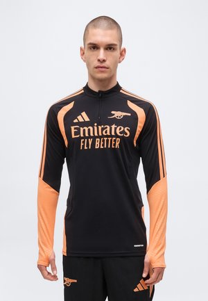 Muž nosící černo-oranžový tréninkový dres Arsenalu s logem Emirates Fly Better a černé šortky s emblémem Arsenalu.