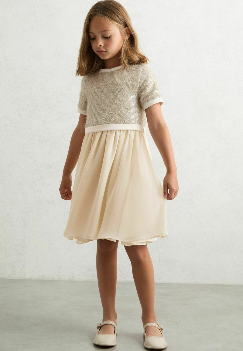 Vestido crema que presenta un top de punto texturizado de mangas cortas y una falda fluida. La falda es plisada, con una tela suave y ligera.
