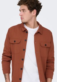 Νεαρός με σγουρά μαλλιά που φοράει μπουφάν-πουκάμισο χρώματος σκουριάς πάνω από λευκό μπλουζάκι, κοιτάζοντας προς τα αριστερά.