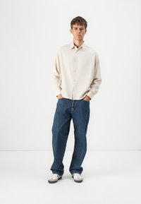 Beige button-up shirt with a textured finish, paired with loose-fitting blue denim jeans and white sneakers. Simple, casual outfit.