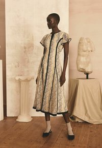 Kleid mit Blumenmuster in Creme und Schwarz, mit kurzen Ärmeln, einem modesten Ausschnitt und einer ausgestellten Silhouette, ergänzt durch schwarze Schuhe und Socken.