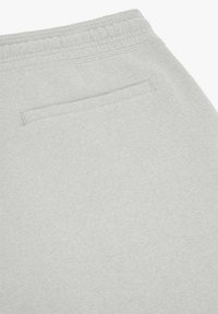 Section d'un pantalon en tissu gris clair montrant une ceinture élastique et une unique poche arrière horizontale avec des coutures discrètes.