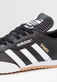 Černé kožené tenisky Adidas Samba s bílými pruhy, texturovanou gumovou podrážkou a zlatým logem "Samba". Ploché černé tkaničky a polstrovaný jazyk.