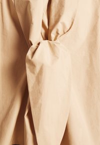 Tissu beige rassemblé et noué fermement au centre, créant des plis et des creux avec une texture lisse.