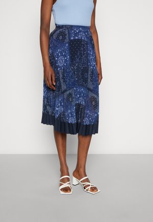 Woman wearing a blue patchwork-patterned pleated midi skirt, light blue sleeveless top, and white strappy block-heeled sandals.