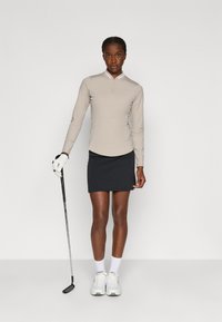Maglione beige a maniche lunghe da golf con colletto a mezza zip, abbinato a una gonna da golf nera. Scarpe da ginnastica bianche e un bastone da golf tenuto nella mano sinistra.