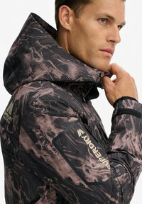Man die de kraag van een zwart en bruin gestippeld Superdry-jack met capuchon en merkemblemen op de mouw en achterkant aanpast.