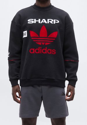 Mann trägt schwarzen Sweatshirt mit großem rotem Adidas-Logo, weißem "SHARP" Schriftzug und grauer Adidas-Shorts und steht vor hellem Hintergrund.
