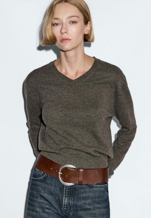 Žena s krátkými blond vlasy, oblečená v tmavě šedém svetr s výstřihem do V, modrých džínách a širokým hnědým koženým opaskem se stříbrnou přezkou, stojící u bílé zdi.