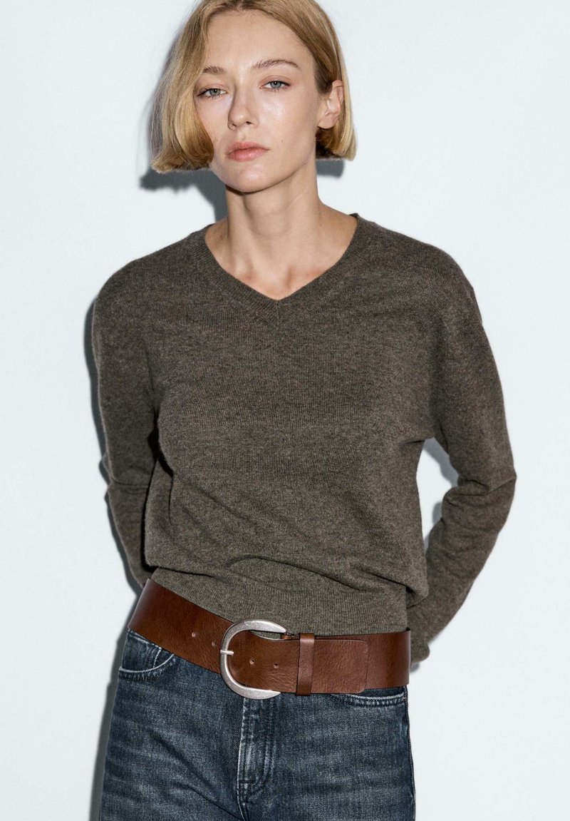 Žena s krátkými blond vlasy, oblečená v tmavě šedém svetr s výstřihem do V, modrých džínách a širokým hnědým koženým opaskem se stříbrnou přezkou, stojící u bílé zdi.