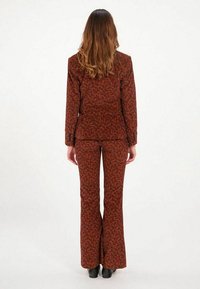 Costume à imprimé floral marron avec une veste ajustée et un pantalon évasé. Tissu texturé avec des détails subtils. Modèle se tient de dos.