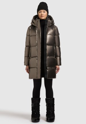 Metallic Puffer-Jacke mit Kapuze, Reißverschluss vorne und gepufften Steppabschnitten. Getragen über einem schwarzen Outfit, kombiniert mit schwarzen Schneestiefeln.