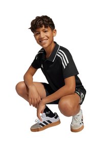 Maglietta polo nera con strisce bianche, pantaloni corti neri e scarpe sportive bicolore con suola color gomma. Capelli castani corti, in posa accovacciata, sorridente.