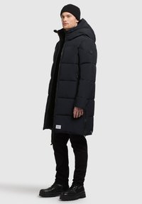 Długi czarny pikowany płaszcz z kapturem, zapinany na zamek, z przednimi kieszeniami i teksturowanym wykończeniem. Noszony z czarnymi spodniami i solidnymi czarnymi butami.