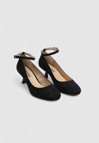 Talons en daim noir avec un bout rond, un petit talon carré et une bride à la cheville. L'intérieur est beige avec une marque discrète. Texture lisse.