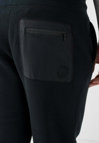 Pantalones deportivos negros con un bolsillo con cremallera y un parche con logo. Tejido suave y texturizado con un corte ajustado y cinturilla elástica.