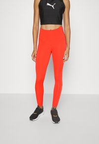 Czerwone legginsy z wysokim stanem, zestawione z czarnym krótkim topem na ramiączkach, na którym znajduje się biały logo. Czarne buty sportowe z białymi akcentami.