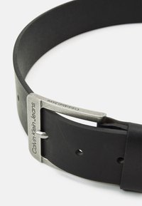 Sort læderbælte med en glat overflade, prydet med en sølvfarvet metalspænde. Spændet har præget branding: "Calvin Klein Jeans."