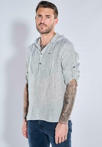 Leichte, gestreifte Langarmbluse in Weiß und Grün mit Kapuze, knöpfbarem Vorderteil und hochgekrempelten Ärmeln. Jeans sind darunter sichtbar.