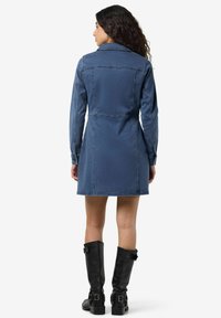 Denimkjole i middels blå med lange ermer, krage og innsvingt midje. Brukt med knelengde svarte lærstøvler.