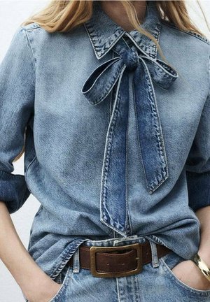 Femme portant une chemise en denim bleu clair avec un grand nœud noué au col, manches retroussées, ceinture en cuir marron avec boucle en laiton et mains dans les poches.