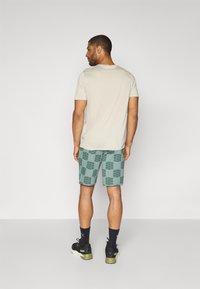 T-shirt de algodão bege, com mangas curtas, combinado com calções teal com um padrão de grelha. Calçado: sapatos pretos com sola verde e meias pretas.