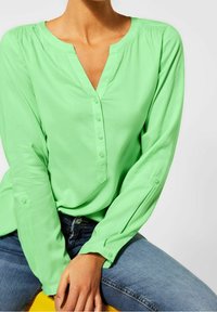 Grön långärmad blus med V-ringning och knappslå, i mjukt tyg och med manschetter, som är ihopparad med jeans i denim.