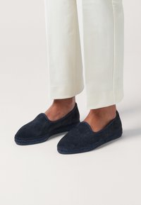 Navy suede slip-on shoes with a smooth finish, rounded toe, and woven side accents, paired with light-colored tailored pants.
