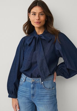 Kobieta o długich brązowych włosach, ubrana w ciemnoniebieską bluzkę z kołnierzem w formie kokardy oraz jasnoniebieskie dżinsy, stojąca z jedną ręką na biodrze.