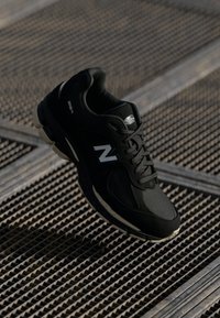 Black New Balance sneaker with white "N" logo and gray mesh details resting on a metal grid surface with shadow patterns.