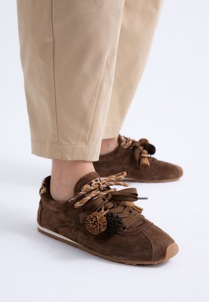 Zapatillas de ante marrón con cordones marrones y estampado de leopardo y detalles de pompón, usadas con pantalones beige recortados sobre un fondo blanco.