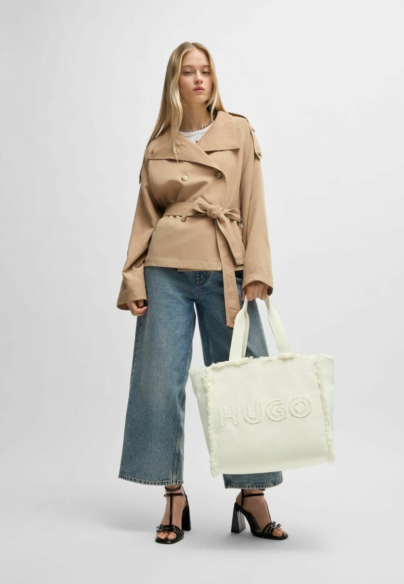 Trench beige con cintura, jeans a gamba larga blu, tacchi neri con cinturini e borsa a mano bianca con logo "HUGO" in rilievo.