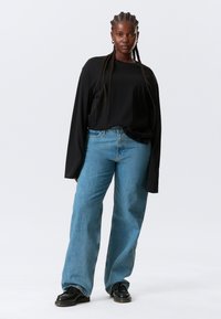 Camisa negra de manga larga, de corte holgado, combinada con jeans azul claro de talle alto. Zapatos negros con cordones y suela gruesa.