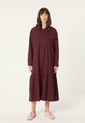 Vrouw staand, een lange mouwen bordeaux midi-jurk met een gerimpelde zoom en borstzakken, gecombineerd met lichtroze sneakers.