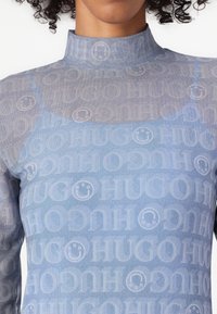 Top ajusté bleu clair avec col montant, présentant un motif répétitif "HUGO" et des visages souriants sur toute la surface, en matériau transparent.