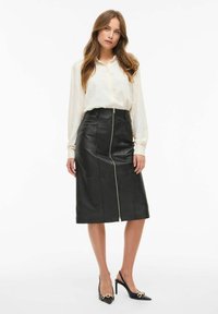 Mustahiilinen musta nahkainen midihame etuvetoketjulla, yhdistettynä kevyesti teksturoituun paitaan ja mustiin korkeakantaisiin slingback-kenkiin, joissa on kultaisia yksityiskohtia.