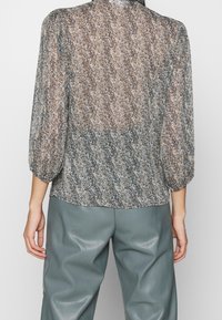 Blusa transparente con un patrón texturizado en tonos de marrón y azul, mangas abullonadas y dobladillo redondeado, combinada con pantalones de cuero sintético grises.