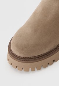 Bottine en suède beige avec un bout arrondi, des coutures marron contrastantes, et une semelle en caoutchouc texturée épaisse avec un motif dentelé.