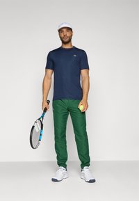 Cămașă sport cu mânecă scurtă de culoare bleumarin, pantaloni sport verzi, șapcă albă, ținând o rachetă de tenis și o minge, adidași albi, stofă netedă, design ajustat.