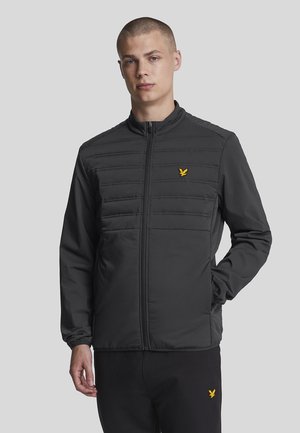 Svart zip-up-jacka med quiltade frontpaneler, hög krage och gult logo på vänster bröst. Slät textur, långa ärmar och figursydd design.