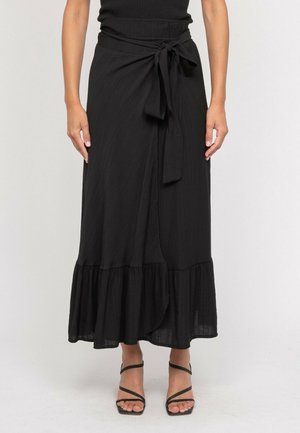 Femme portant une jupe portefeuille noire avec un nœud noué à la taille et un volant à l'ourlet, associée à des sandales noires à lanières.