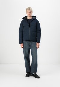 Navy puffer jacket with a hood, featuring a zip front, horizontal quilted design, and paired with straight-leg denim jeans and black shoes.